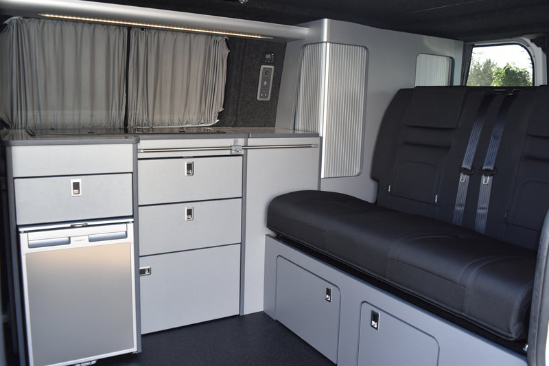 Compact living space with a bench, drawers, and kitchenette inside a vehicle.