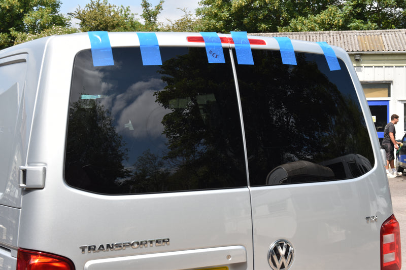 Back of a white Volkswagen van with blue tape on the windows, parked outdoors.
