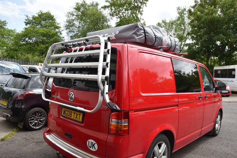 Red van with a luggage rack and roof storage on a street.